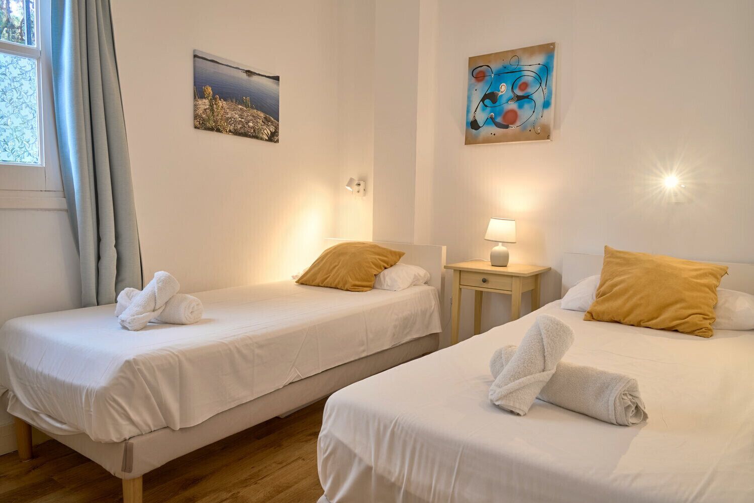 Dos camas individuales con sábanas blancas y almohadas amarillas, una mesita de noche con lámpara y una ventana con cortinas.