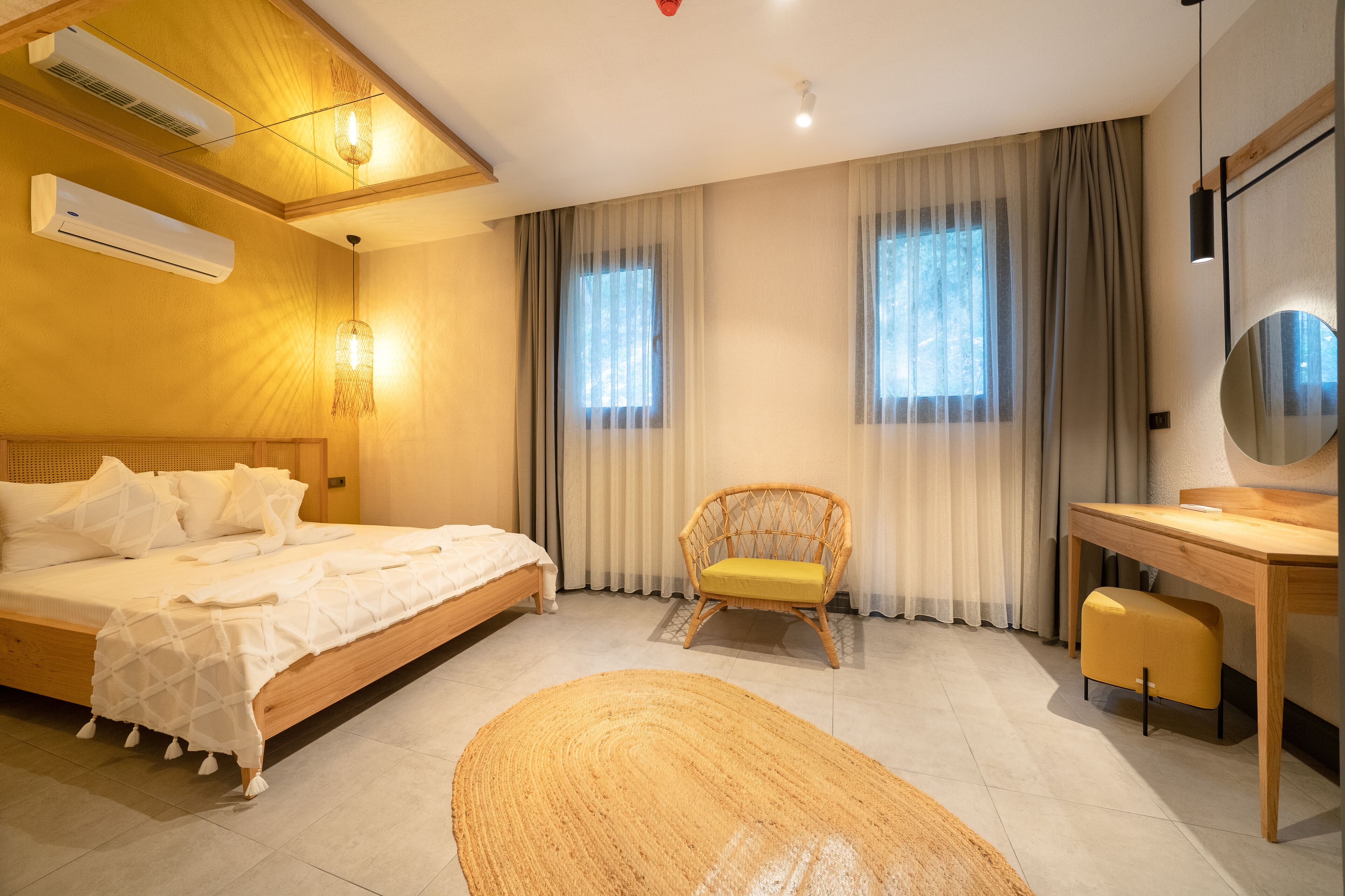 A modern hotel room with a wooden bed, a wicker chair, a desk, and large windows with sheer curtains.