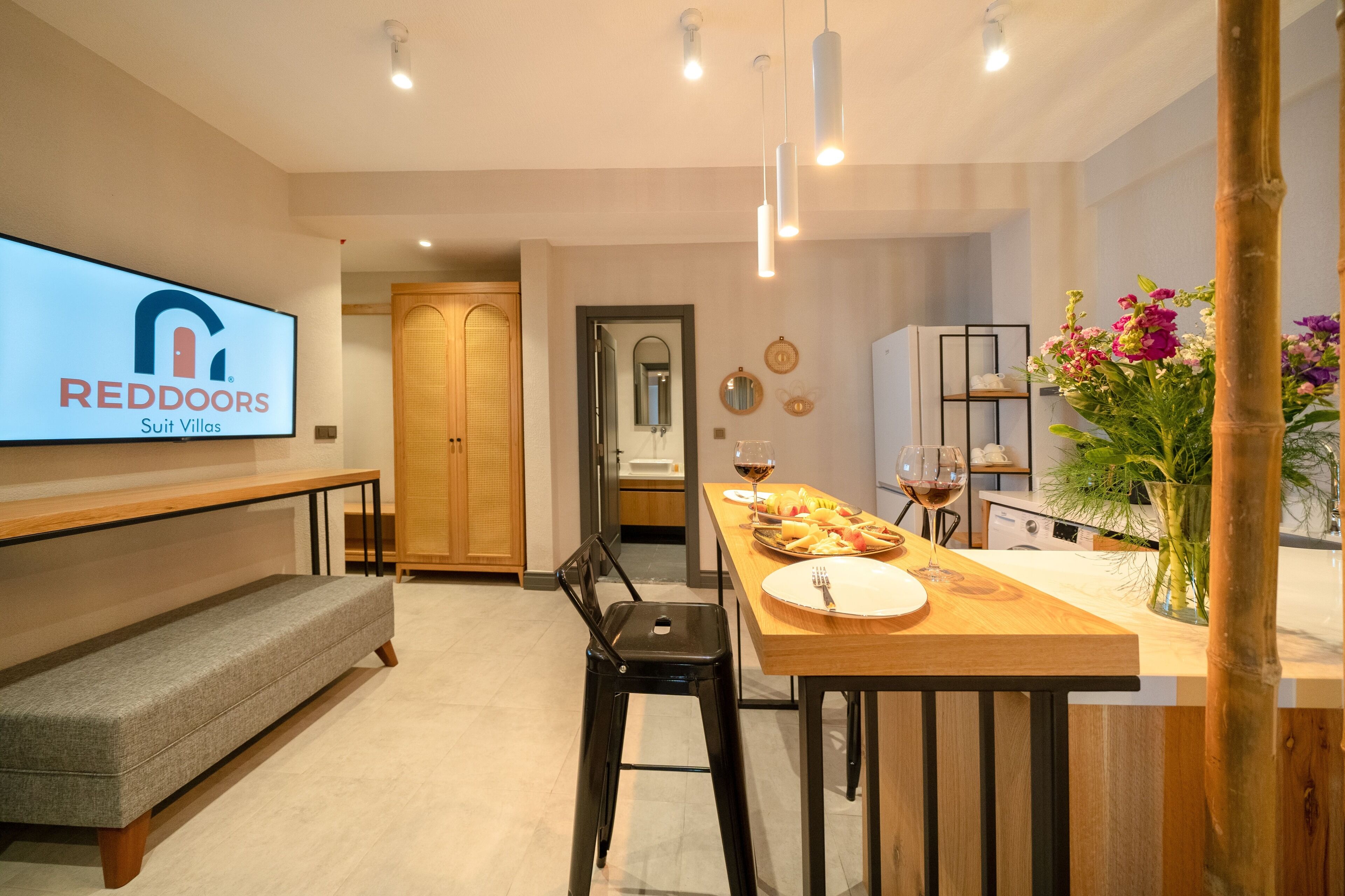 A modern kitchen with a wooden dining table, bar stools, and a TV displaying 'REDDOORS Suit Villas'.