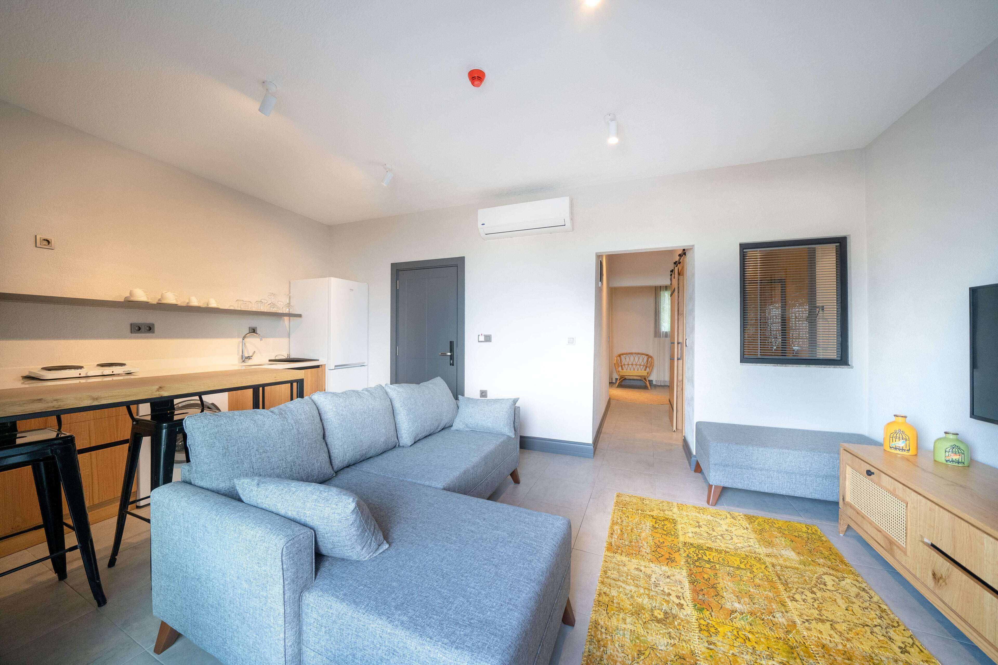 A modern living room with a grey sectional sofa, a wooden dining table, and a yellow patterned rug.