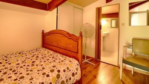 A bedroom with a wooden bed, a polka dot bedspread, a standing fan, and a chair.