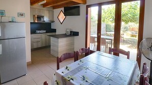 A compact kitchen with a refrigerator, microwave, and coffee maker. There is a dining table with chairs and a fan. The kitchen opens to a patio with outdoor furniture.