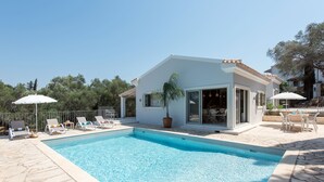 A swimming pool with lounge chairs and umbrellas, surrounded by a white building with a tiled roof. - Villa Helonaki (CORFU)