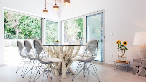 A modern dining area with a glass table surrounded by wire chairs, large windows, and a view of greenery outside. - Villa Helonaki (CORFU)