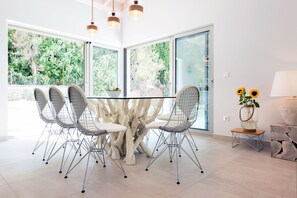 A modern dining area with a glass table surrounded by wire chairs, large windows, and a view of greenery outside. - Villa Helonaki (CORFU)