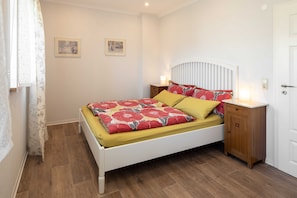 A bedroom with a white bed, yellow and red floral bedding, two wall-mounted pictures, a bedside table with a lamp, and a wooden nightstand.