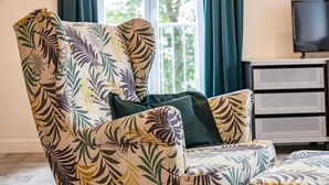 A room with a large armchair and ottoman featuring a tropical leaf pattern. A television is mounted on the wall, and there is a window with curtains. - Mauersegler - Old Castle Brewery Apartments (Bad Wildungen)