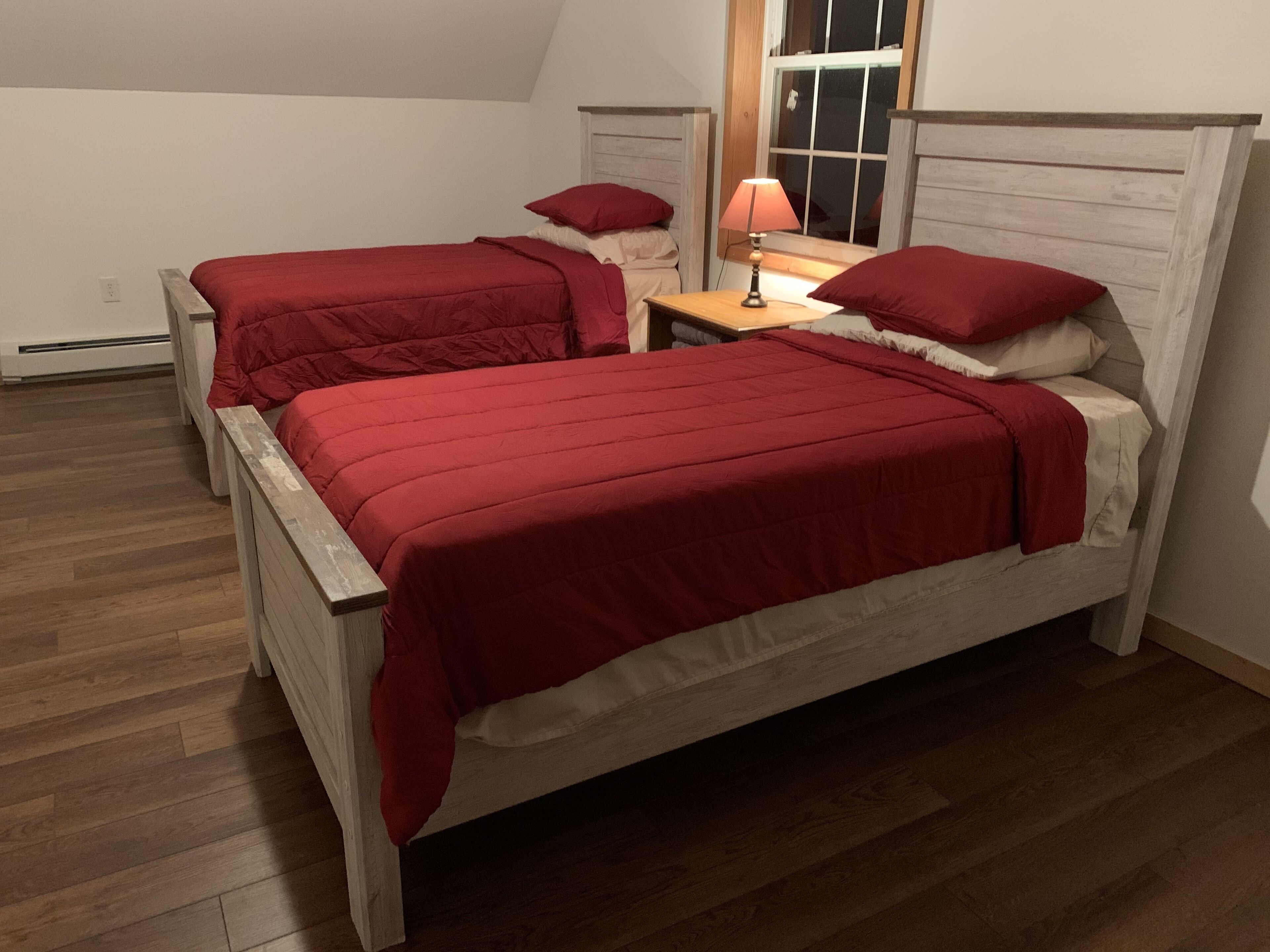 Quarto com duas camas, cada uma com roupa de cama vermelha, uma mesa de cabeceira com abajur e uma janela com persianas de madeira.