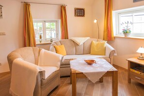 Ein gemütliches Wohnzimmer mit einer Couch, Sesseln, einem Tisch mit einer Schale, einer Lampe und einem Fenster mit Vorhängen.