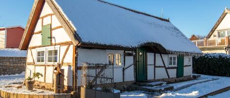 A traditional half-timbered house with a snow-covered roof, a green door, and a small garden area with potted plants.