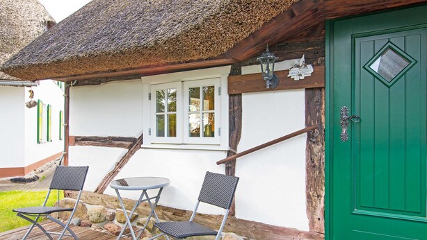 Un edificio rústico con puerta verde, techo de paja y un pequeño espacio al aire libre con dos sillas y una mesa.