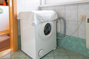 A compact, white washing machine with a hose attached, situated in a room with green-tiled walls and a wooden door frame.