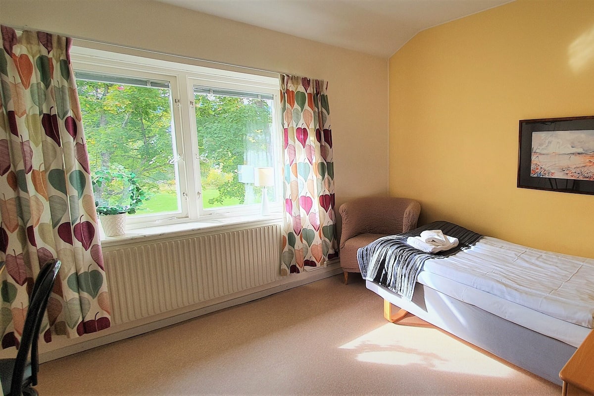 A small, well-lit bedroom with a single bed, a chair, a bedside table with a lamp, and a window with curtains.