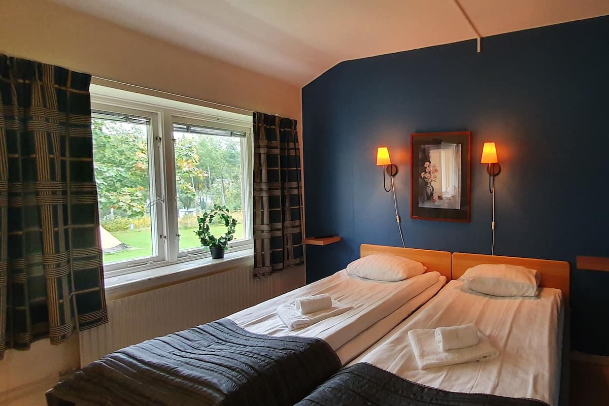 A hotel room with two beds, a window with curtains, a wall-mounted lamp, and a framed picture.