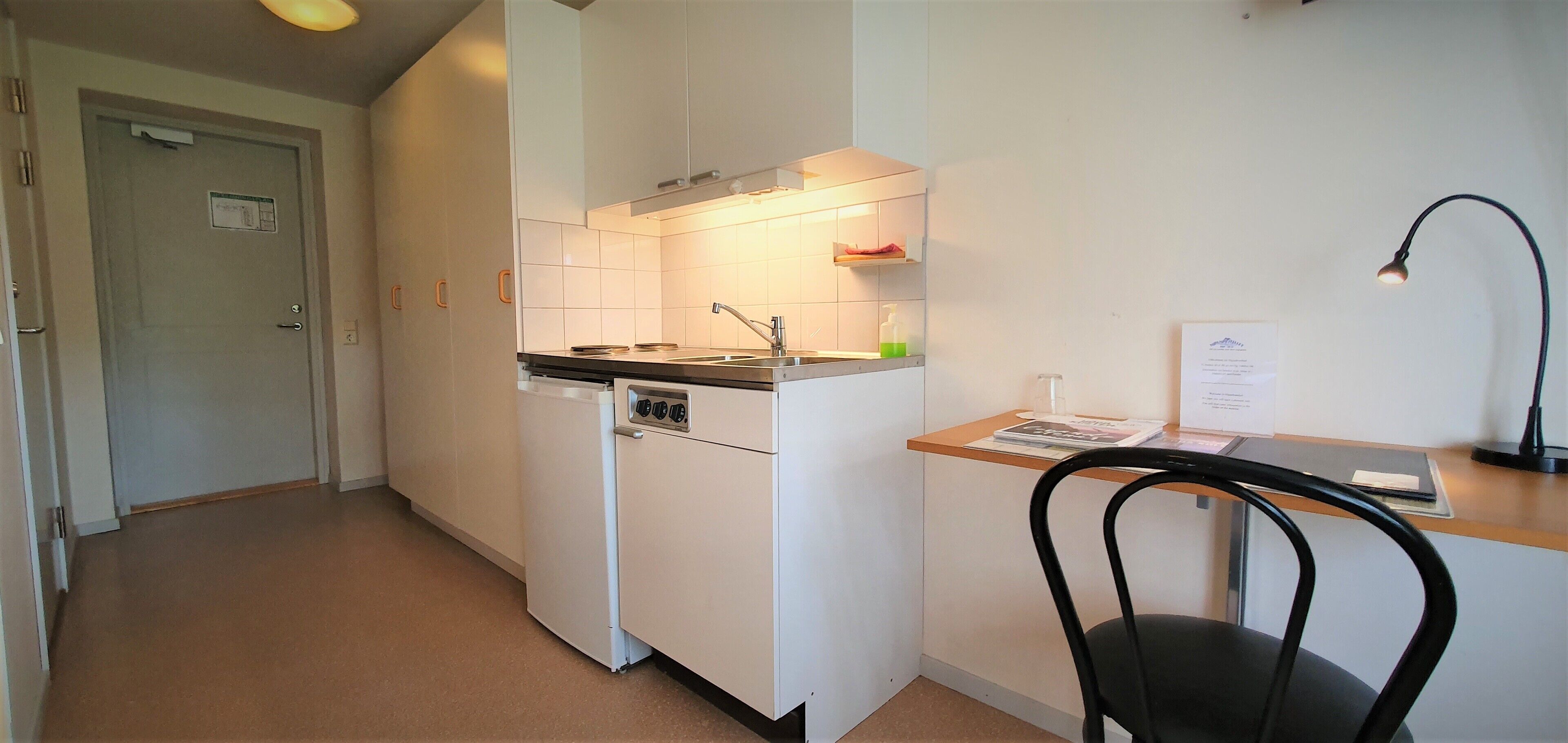 Cocina compacta equipada con refrigerador, estufa y lavaplatos. Incluye una silla y un escritorio con lámpara.