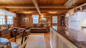 L’intérieur d’un chalet rustique présente un plafond en bois, un espace de vie chaleureux avec un canapé en cuir, une table à manger et un coin cuisine équipé d’un réfrigérateur et de meubles de rangement en bois.