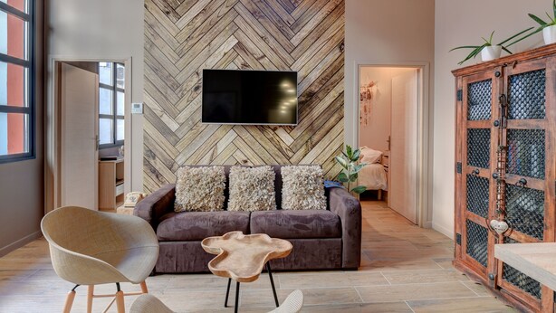 Een moderne woonkamer met een lappendeken patroon aan de muur, een flatscreen televisie, een bruine bank met decoratieve kussens, een bijzondere houten salontafel en een rustieke houten kast met planten.