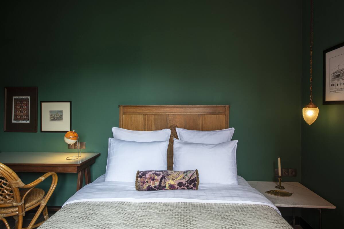 A neatly made bed with white linens and a wooden headboard, a wicker chair, a small table with a lamp, and framed pictures on the wall.
