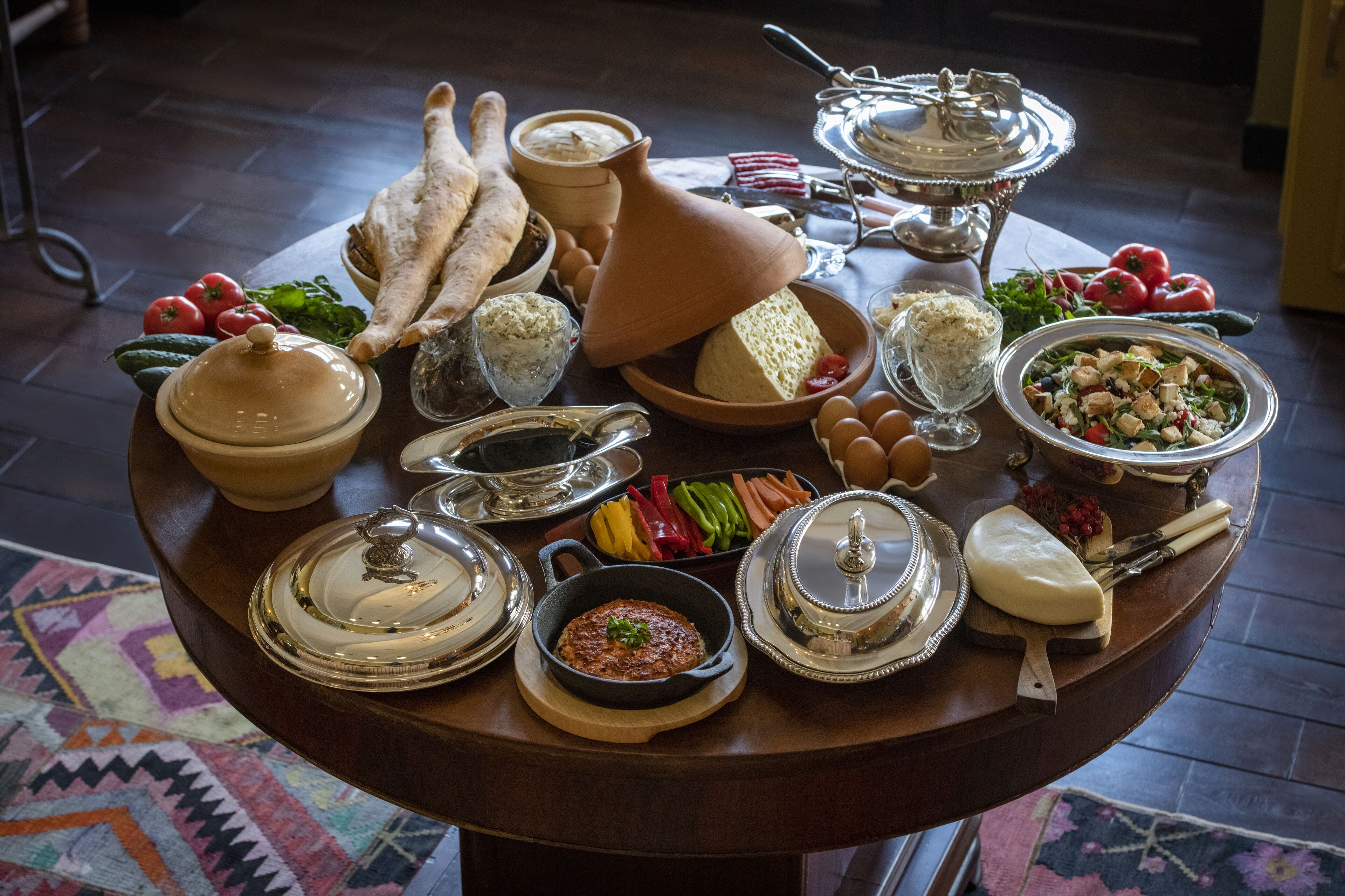 Ein runder Holztisch mit verschiedenen Speisen und Besteck, darunter Käse, Brot und Gemüse.