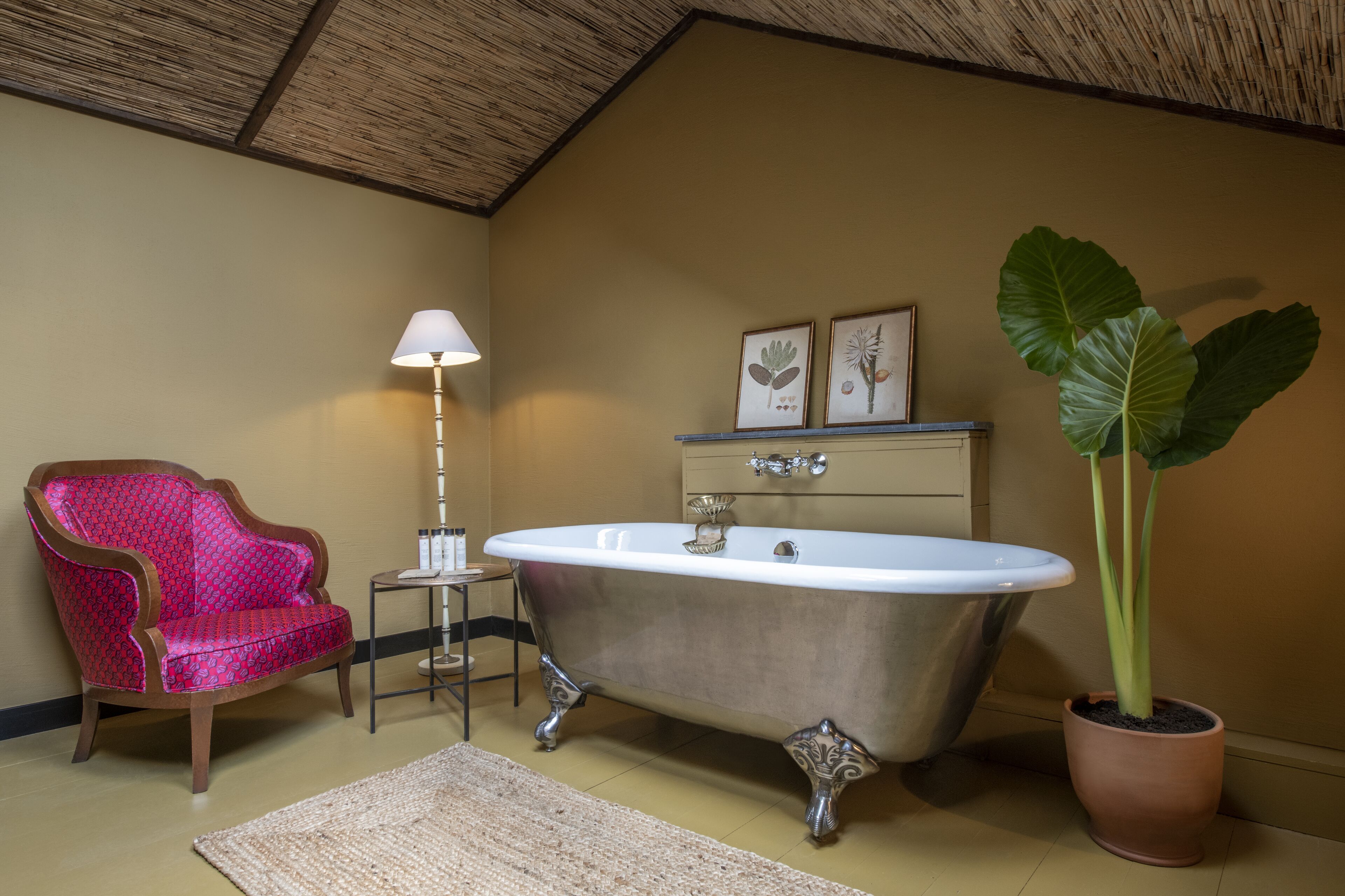 A bathroom with a clawfoot bathtub, a red patterned chair, a small side table, a floor lamp, and a large potted plant.