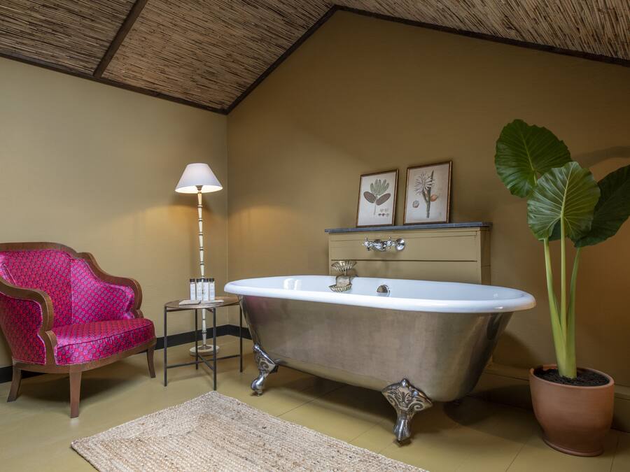 Un baño con una bañera de patas de león, una silla roja con estampado, una mesita auxiliar, una lámpara de pie y una planta grande en maceta.