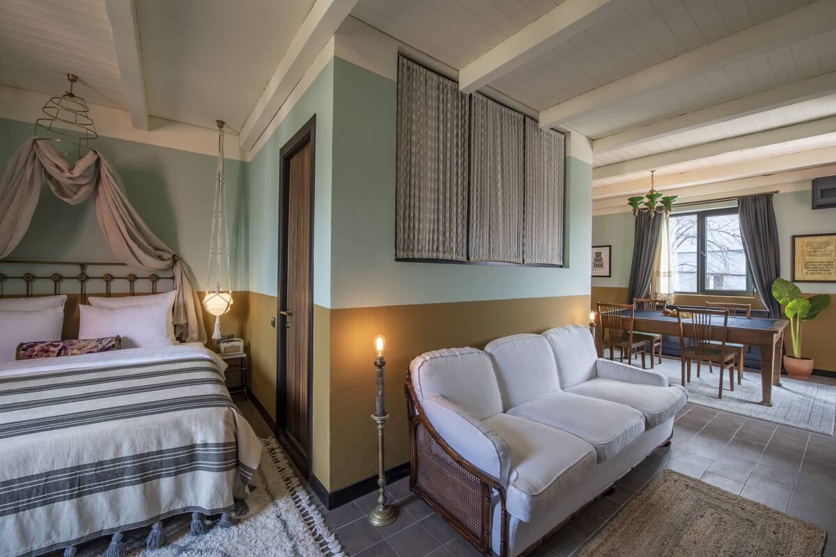 A bedroom with a bed, a nightstand, a lamp, a sofa, a dining table with chairs, and a window with curtains.