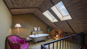 Una habitación con techo inclinado, bañera, una silla, una lámpara y una ventana.