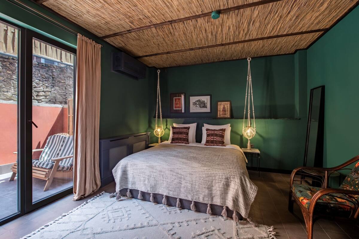 A bedroom with a large bed, a hanging light fixture, a chair, and a view of the outside.