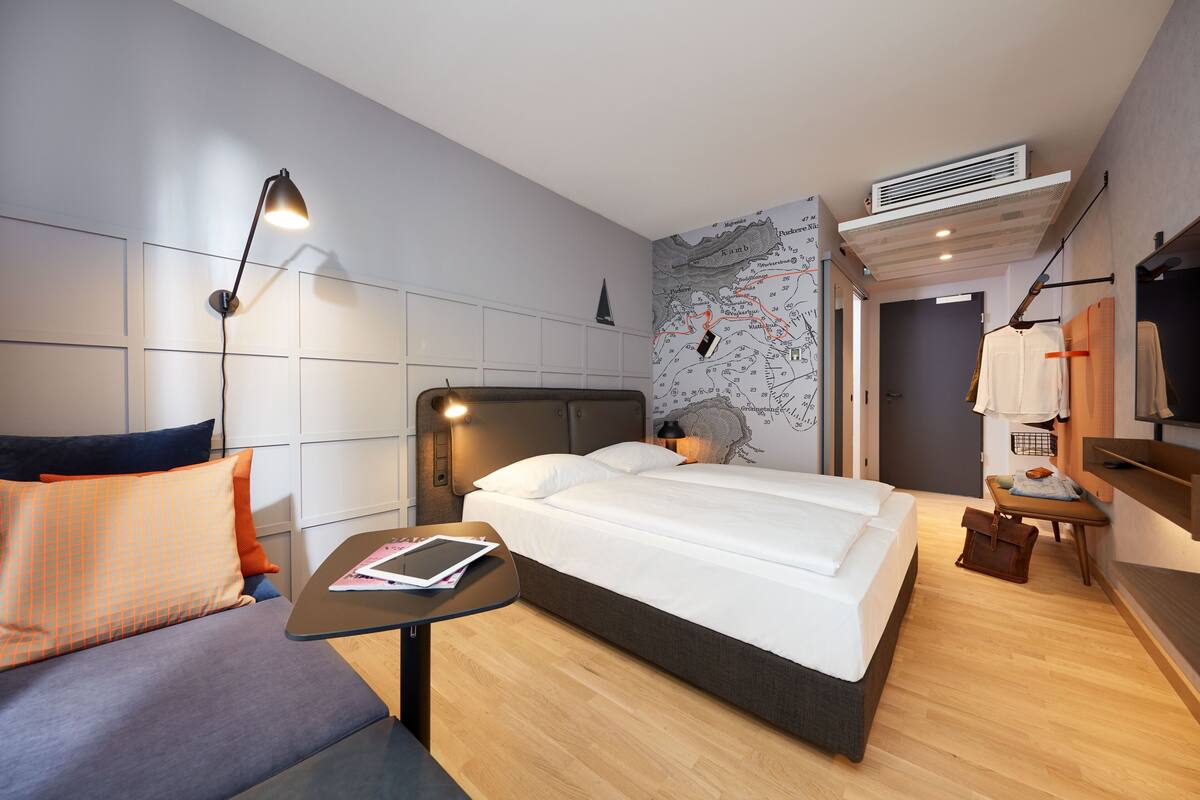 Ein modernes Hotelzimmer mit einem Bett, einer Couch, einem kleinen Tisch mit einem Buch, einer Karte an der Wand und einem Fernseher auf einem Schrank.