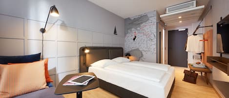 Una habitación de hotel moderna con cama, sofá, una mesita con un libro, un mapa en la pared y televisor sobre un mueble.