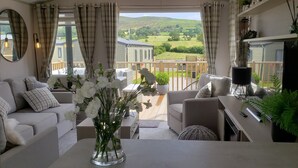 A cozy living room with a sofa, armchair, and a table with a vase of flowers. A balcony with a wooden railing and a view of green hills is visible through the window. - Beautiful 2-bed Lodge Ribble Valley Clitheroe (Clitheroe)