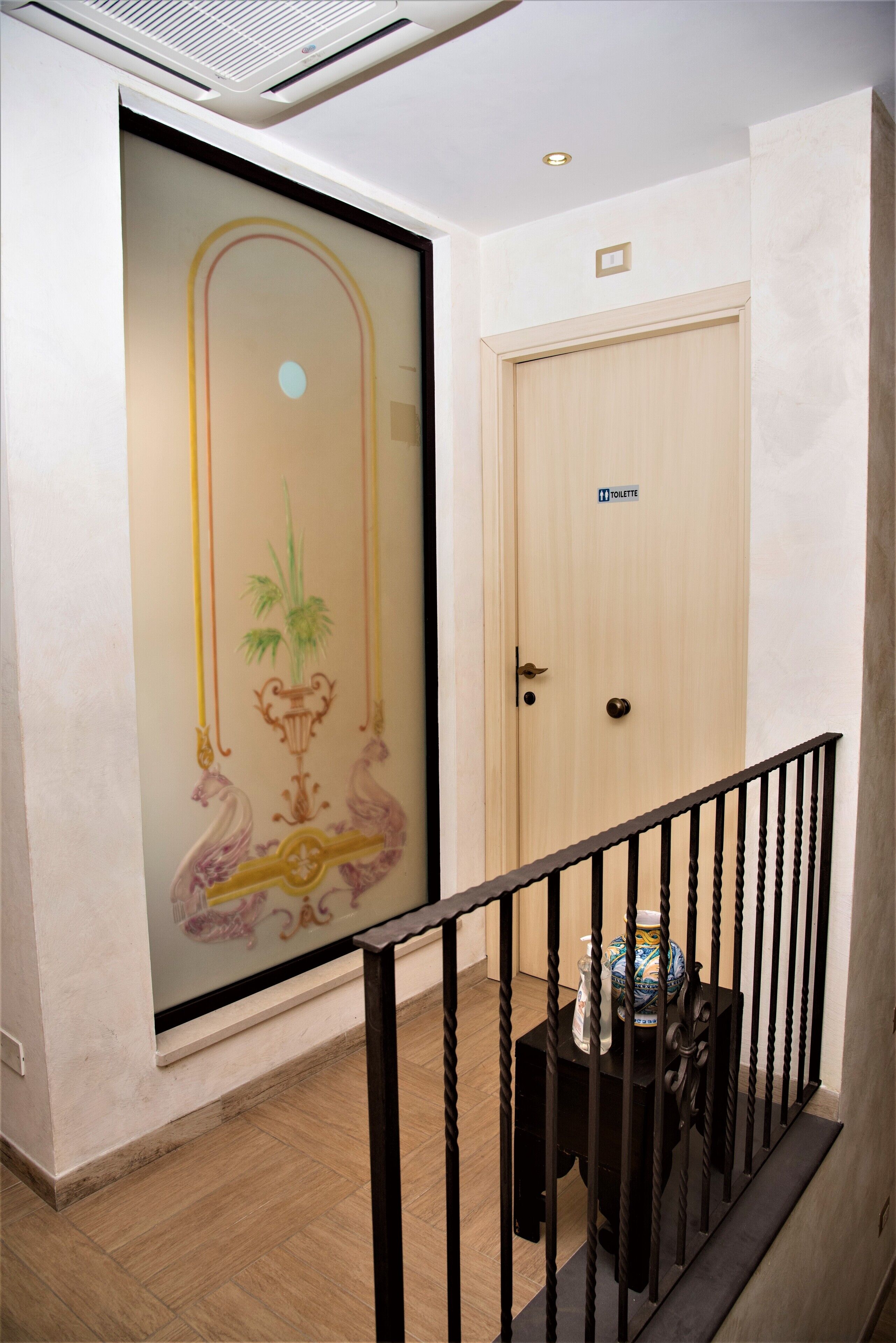 A hallway with a wooden door labeled "Toilette," a glass door with decorative patterns, and a small table with a vase and a bowl.