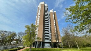 A tall, modern building with a unique curved design, surrounded by green trees and a clear sky.