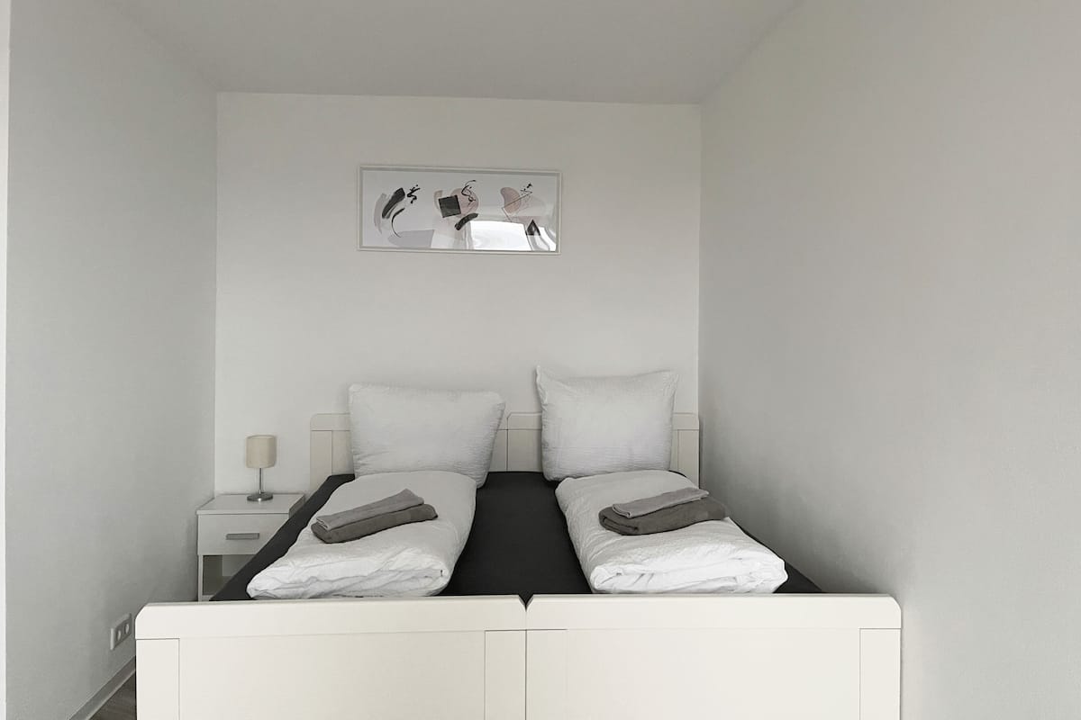 Een kleine, witte eenpersoonskamer met een nachtkastje, een lamp en een ingelijst plaatje aan de muur.