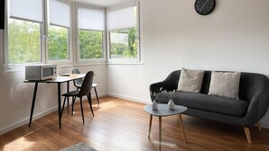 A modern living room with a grey sofa, a small round table, a dining table with chairs, a microwave, and a clock on the wall.