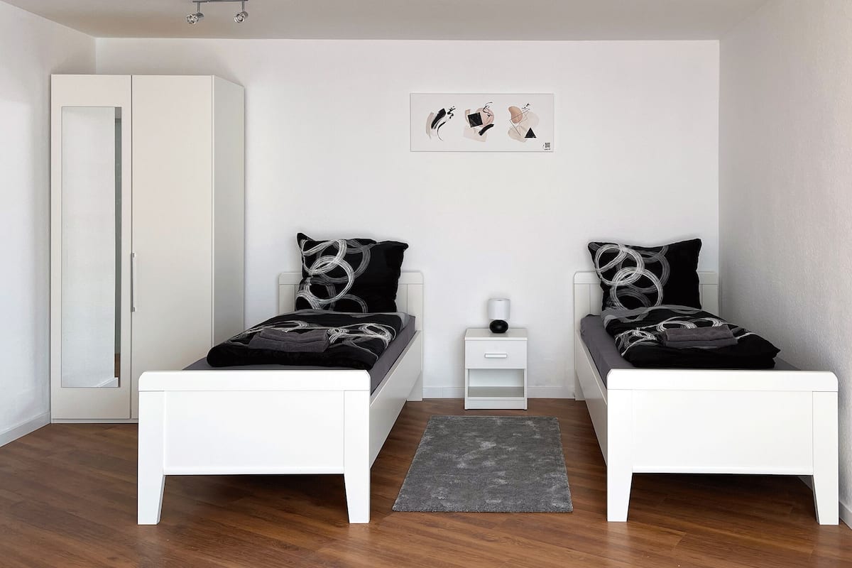 Een minimalistische slaapkamer met twee bedden, een nachtkastje, een grijs vloerkleed en een kunstwerk aan de muur.