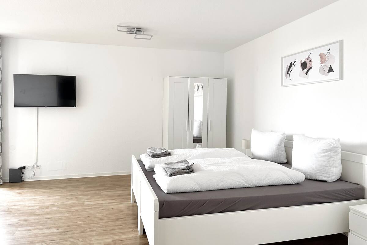 Een moderne slaapkamer met een bed, een aan de muur bevestigde televisie, een kledingkast en een kunstwerk met abstracte vormen.