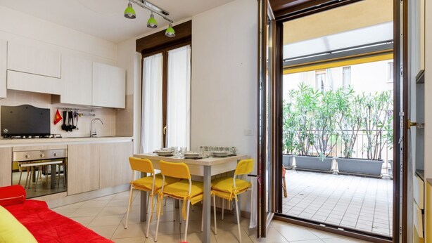 Una cocina moderna con armarios blancos, una mesa de comedor con sillas amarillas y un gran ventanal corredizo que da a un balcón con plantas en macetas.