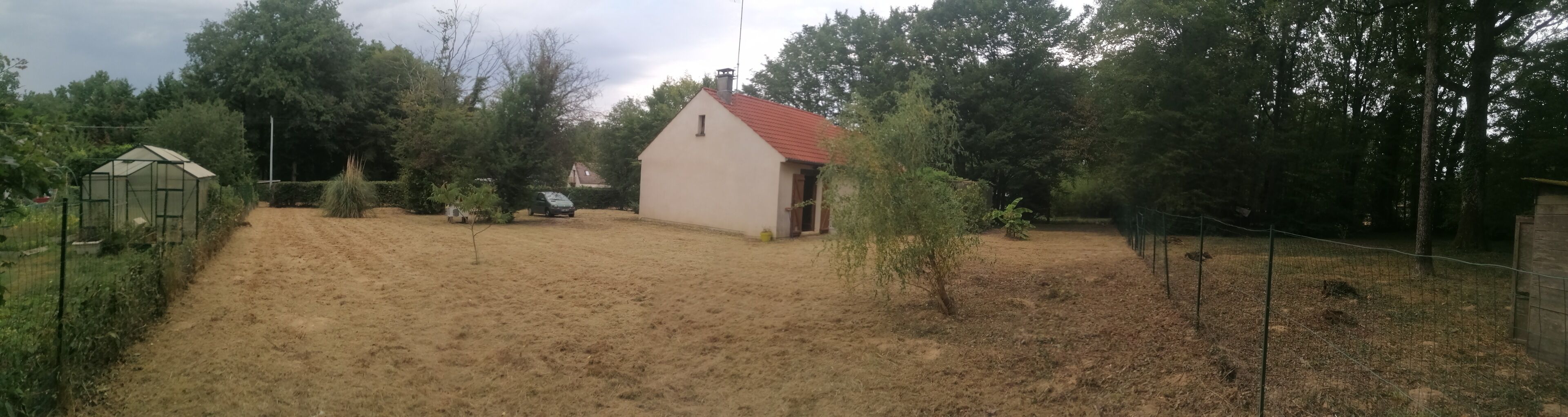 A small, single-story building with a red roof, surrounded by a fenced yard with sparse grass and a few trees.