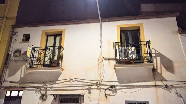 En bygning med to balkoner, hver med gelænder og et lille vindue. Synlige elledninger og en lygtepæl er placeret overfor.