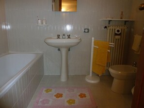 Ein Badezimmer mit einem weißen Waschbecken, einer Badewanne und einer Toilette. Über dem Waschbecken hängt ein Spiegel und darunter befindet sich ein Ablagefach mit Pflegeprodukten.