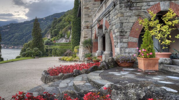 Ein historisches Gebäude mit Garten, Kiesweg und einem See mit Bergen im Hintergrund.
