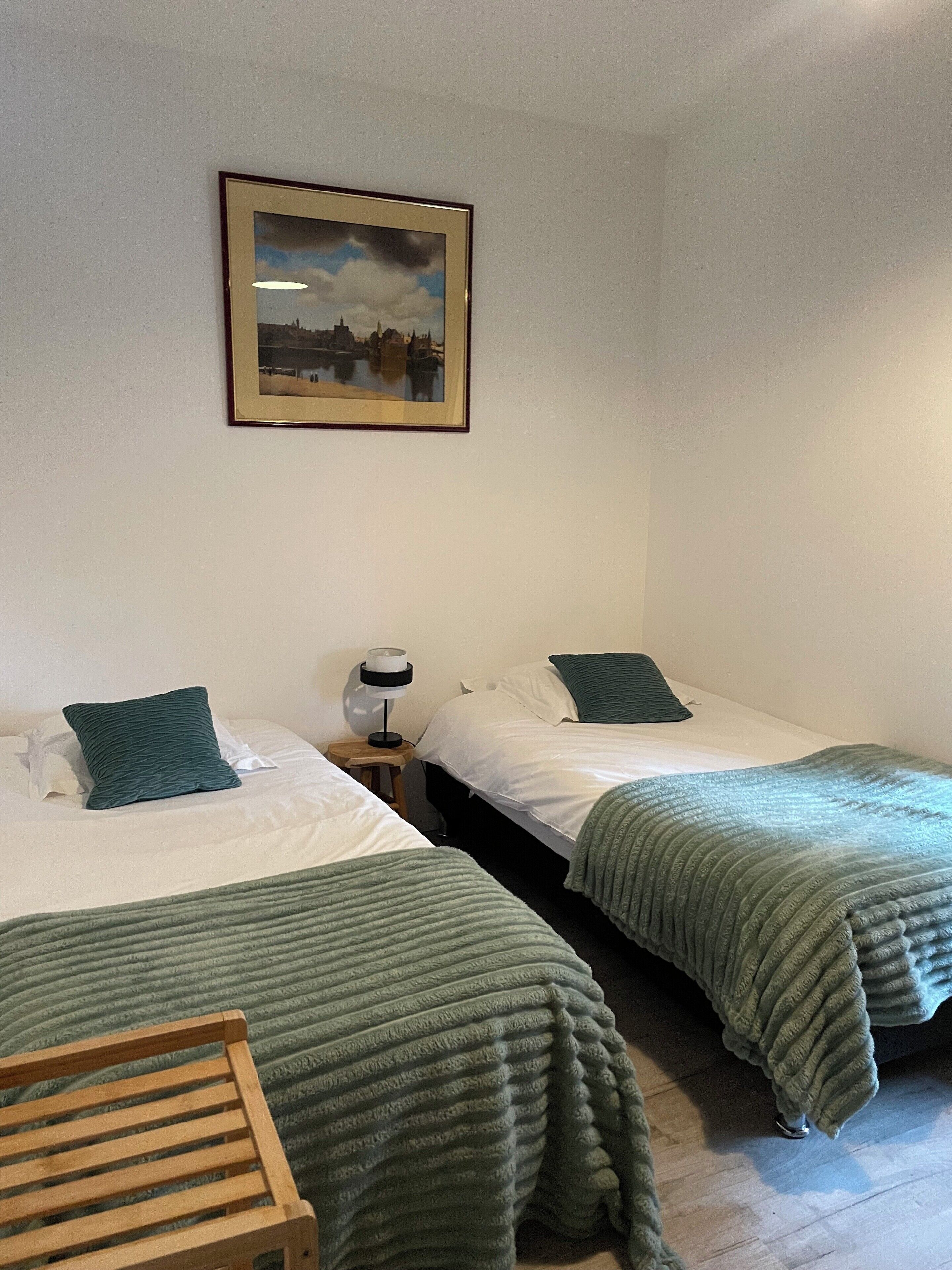 A room with two beds, each with a green blanket and pillow, a small bedside table with a lamp, and a framed picture on the wall.