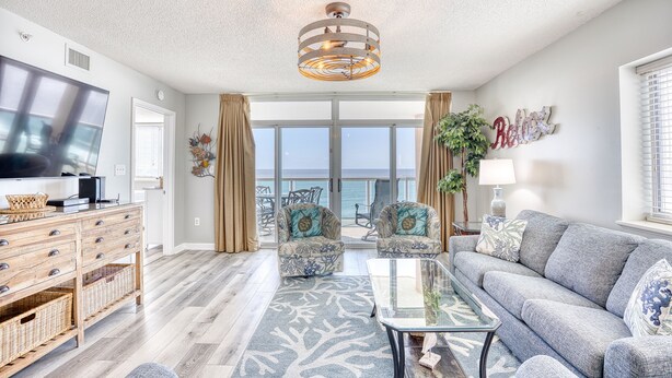 A spacious living room with a large flat-screen TV, a grey sofa, two patterned armchairs, a glass coffee table, and a ceiling light fixture. - Crescent Keyes 810 | Oceanfront 3 Bedroom Condo (North Myrtle Beach)