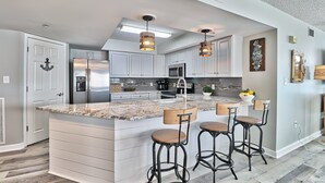 A modern kitchen with a large island, bar stools, stainless steel appliances, and pendant lighting. - Crescent Keyes 810 | Oceanfront 3 Bedroom Condo (North Myrtle Beach)