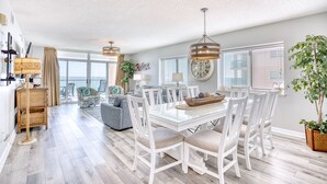 A spacious dining area with a glass table and white chairs, a wicker side table, and a large window with blinds. - Crescent Keyes 810 | Oceanfront 3 Bedroom Condo (North Myrtle Beach)