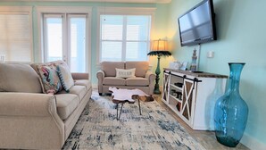 Ein Wohnzimmer mit einem beigen Sofa, einem kleineren Sofa mit einem 'Beach Bum'-Kissen, einem Couchtisch, einer blauen Vase und einem an der Wand montierten Fernseher.