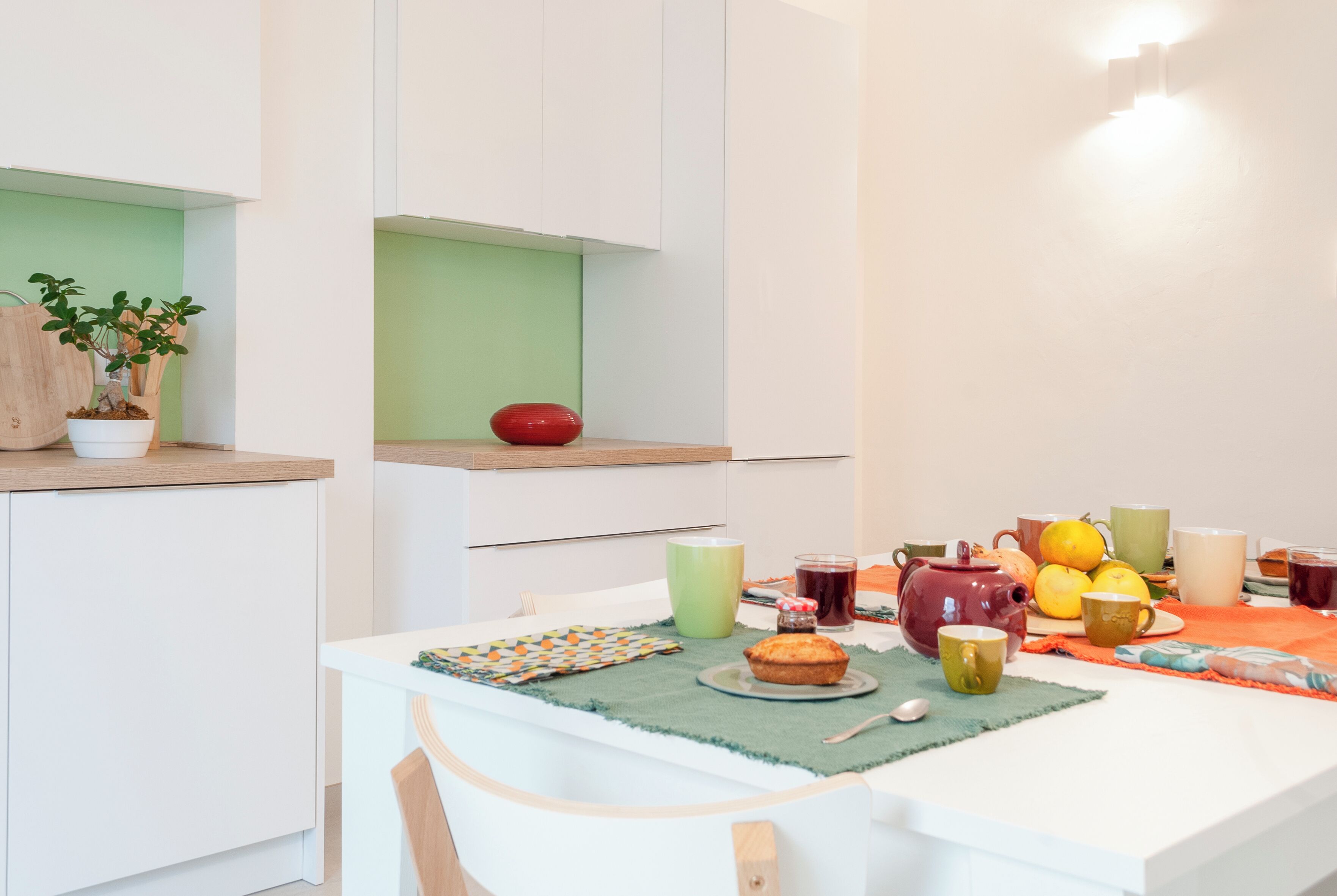 Eine moderne Küche mit weißen Schränken, einer grünen Arbeitsplatte und einem Esstisch, gedeckt mit einem Muffin, Tassen und einem Teekessel.