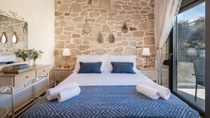 A bedroom with a stone wall, a bed with blue bedding, two pillows, a nightstand with a lamp, and a window with a view of the outside. - Villa Vanka (Chania)