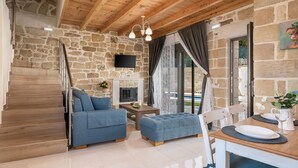 A cozy room with a stone wall, wooden ceiling, a TV, a fireplace, a dining table with chairs, and a staircase. - Villa Vanka (Chania)